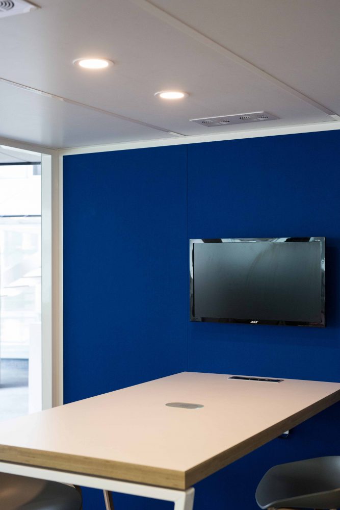 Meeting Box von innen mit Arbeitstisch, Elektroanschlüssen und schwarzem Bildschirm an blauer Rückwand